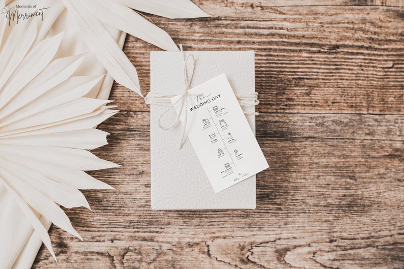 May include: A white gift box tied with twine and a white tag that says "The Wedding Day" with a list of wedding events and times. The box is on a wooden surface with a white palm leaf fan.