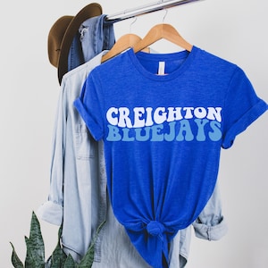 May include: A royal blue t-shirt with a white and blue retro-style graphic that reads "CREIGHTON BLUEJAYS".