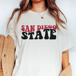 May include: White t-shirt with a red and black graphic that says "SAN DIEGO STATE" in a retro font.