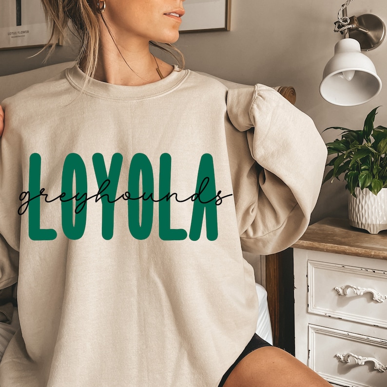 Pode incluir: Uma camisa de moletom bege com o texto "LOYOLA" em letras grandes verdes e o texto "greyhounds" em escrita cursiva abaixo.