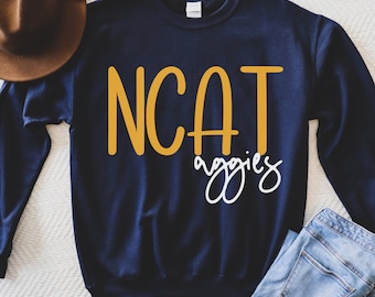 Peut inclure: Un sweat-shirt bleu marine avec le texte "NCAT Aggies" en lettres jaunes et blanches.