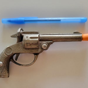 Op de afbeelding: Een vintage speelgoedpistool van zilverkleurig metaal met een oranje punt. Een blauwe pen is boven het speelgoedpistool geplaatst. Het speelgoedpistool heeft een gedetailleerde handgreep en trekker.