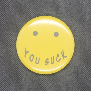 May include: A yellow button with a smiley face design. The button has two gray dots for eyes and the words "YOU SUCK" in gray text. The button is round and has a glossy finish.