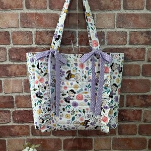 May include: A white tote bag with a floral and cartoon character print. The bag features purple polka dot bows and straps. The bag has a front pocket and is hanging against a brick wall.