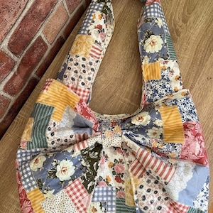 May include: A patchwork tote bag with a bow detail. The bag features a variety of floral and patterned fabrics in shades of blue, yellow, red, and white. The handles are made of the same patchwork fabric.