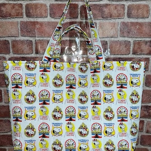 May include: White tote bag featuring a repeating pattern of Snoopy in various designs, including a crown and the text "THE PEOPLE'S CHOICE". The bag has matching handles and is against a brick wall.