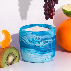 May include: A blue and white marbled candle with the text "LA VIE MARINE Mixed Fruit 4 oz." is surrounded by fresh fruit. The candle has a white wick and a round shape. The fruit includes grapes, an orange, a lime, a kiwi, and a tangerine.