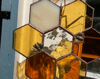 Può includere: Favo in vetro colorato con pannelli esagonali ambra, oro e color crema. Sono attaccati due ciondoli a forma di ape in bronzo. I pannelli sono incorniciati con un metallo color rame. Il pezzo è sospeso da una corda in fibra naturale.