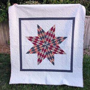 May include: A square quilt featuring a central eight-pointed star pattern composed of small, multi-colored geometric fabric pieces in shades of red, blue, and yellow, surrounded by a dark gray border and a white background with swirling stitching.