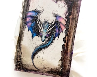 Op de afbeelding: Een fantasie-thema dagboek met een gedetailleerde drakenillustratie. De draak heeft blauwe, paarse en roze vleugels en schubben. Het dagboek heeft een distressed, getextureerde rand en een crème lint.