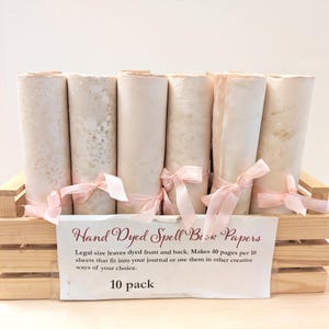 May include: A wooden crate holds several rolled papers tied with pink ribbons. The papers are a light beige color, and the sign in front reads "Hand Dyed Spell Book Papers" and "10 pack".