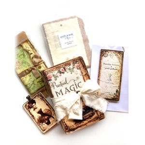 Könnte beinhalten: Eine Auswahl an Zauberbüchern im Vintage-Stil und verwandten Artikeln. Das zentrale Buch mit dem Titel "Practical Magic" ist mit einer cremefarbenen Schleife und floralen Akzenten verziert. Weitere Artikel sind ein Anhänger und ein "Personal Almanac".