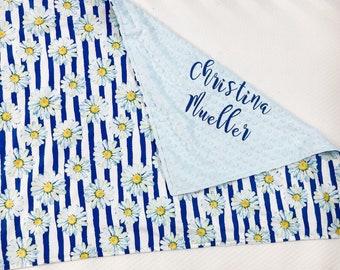 Puede incluir: Una manta para bebé azul y blanca con un estampado de margaritas en un lado y una tela minky azul claro en el otro. La tela minky tiene el nombre "Christina Mueller" bordado en hilo azul.