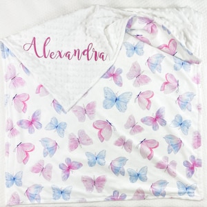 Puede incluir: Una manta blanca para bebé con un patrón de mariposas rosas y azules. La manta tiene un respaldo suave de minky blanco y está personalizada con el nombre "Alexandra" en bordado rosa.