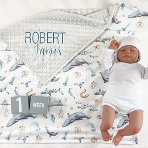 May include: A white baby blanket with a gray and white minky fabric on top. The blanket features a blue and white ocean-themed print with whales, fish, and seahorses. The blanket also has a gray block with the number "1" and the word "week" on it. The blanket is personalized with the name "Robert James".