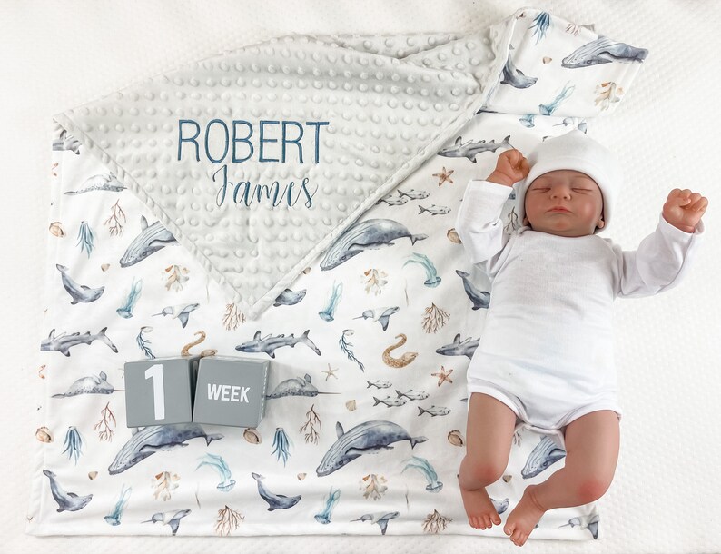 May include: A white baby blanket with a gray and white minky fabric on top. The blanket features a blue and white ocean-themed print with whales, fish, and seahorses. The blanket also has a gray block with the number "1" and the word "week" on it. The blanket is personalized with the name "Robert James".
