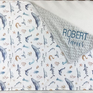May include: A white and gray baby blanket with a blue and brown watercolor ocean print. The blanket has a soft, minky fabric on one side and a cotton fabric on the other. The blanket is personalized with the name "ROBERT James" in blue embroidery.