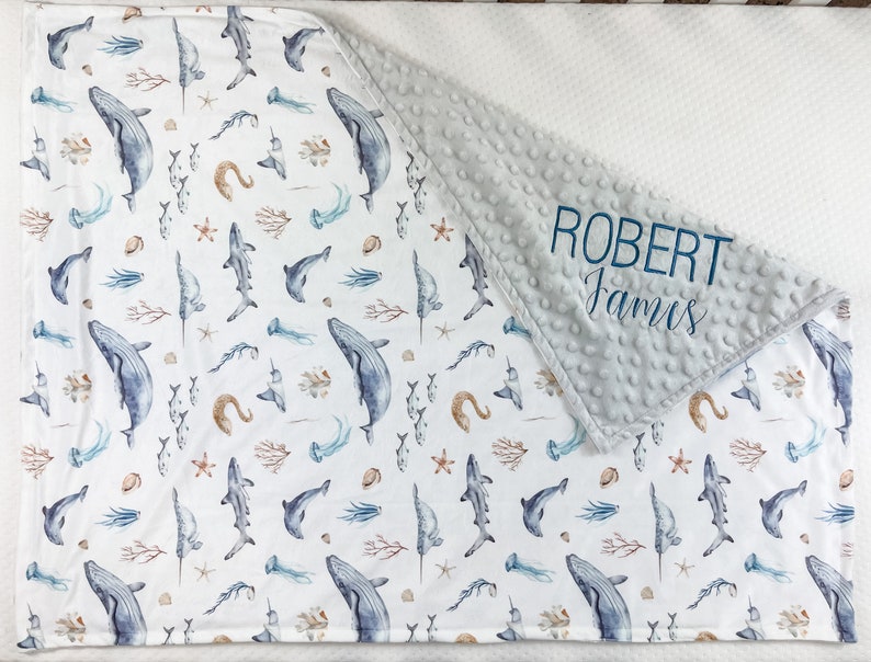 May include: A white and gray baby blanket with a blue and brown watercolor ocean print. The blanket has a soft, minky fabric on one side and a cotton fabric on the other. The blanket is personalized with the name "ROBERT James" in blue embroidery.