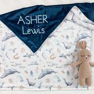May include: A blue and white baby blanket with a sea life pattern. The blanket has a dark blue velvet backing with the name "Asher Lewis" embroidered in light blue thread. A brown crocheted bunny is lying on the blanket.