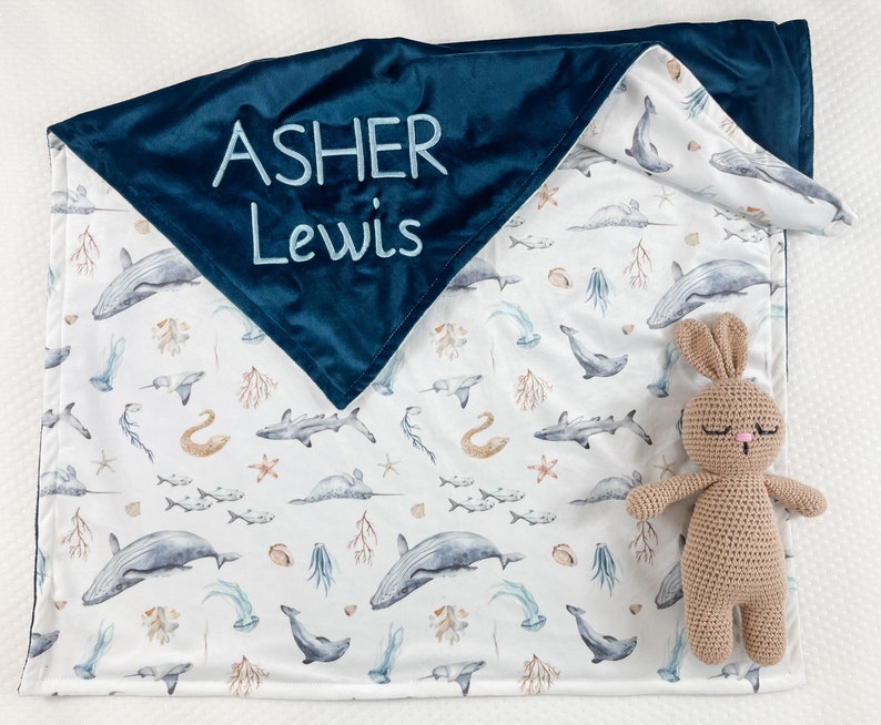 May include: A blue and white baby blanket with a sea life pattern. The blanket has a dark blue velvet backing with the name "Asher Lewis" embroidered in light blue thread. A brown crocheted bunny is lying on the blanket.
