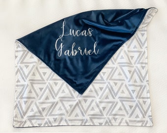 Może przedstawiać: Kocyk dla niemowlęcia w kolorze granatowym i białym z geometrycznym wzorem. Na kocyku wyhaftowany jest napis "Lucas Gabriel" białą nicią.