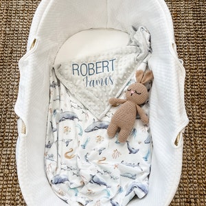 May include: A white wicker bassinet with a gray and white blanket with the name "Robert James" embroidered on it. A brown crocheted bunny is lying on top of the blanket.