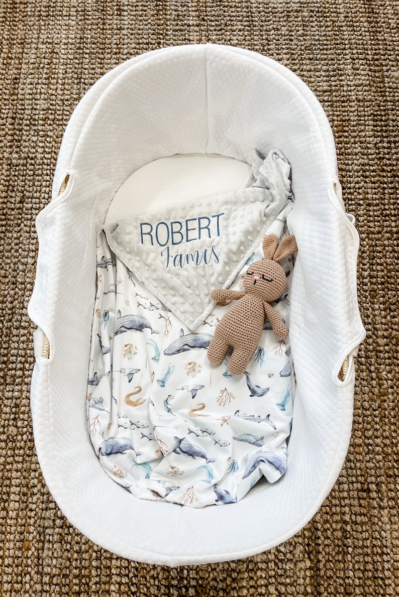 May include: A white wicker bassinet with a gray and white blanket with the name "Robert James" embroidered on it. A brown crocheted bunny is lying on top of the blanket.