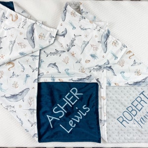 May include: A white baby blanket with a blue and white ocean animal print. The blanket has two personalized squares, one with the name "ASHER LEWIS" in blue embroidery on a dark blue background, and the other with the name "ROBERT JAMES" in gray embroidery on a light gray background.