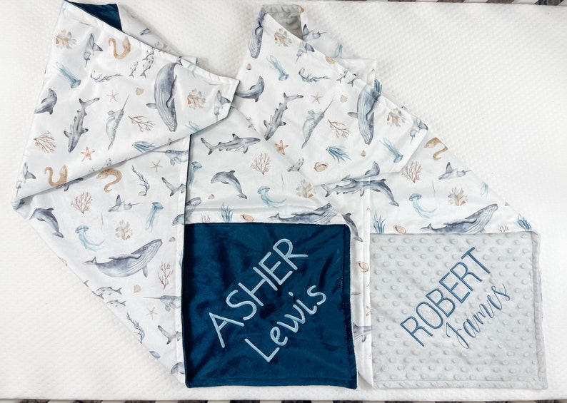 May include: A white baby blanket with a blue and white ocean animal print. The blanket has two personalized squares, one with the name "ASHER LEWIS" in blue embroidery on a dark blue background, and the other with the name "ROBERT JAMES" in gray embroidery on a light gray background.