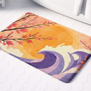 May include: A bath mat with a watercolor design of a sunset over the ocean. The mat features a large orange sun with pink and red branches of a tree on either side. The bottom of the mat features purple and blue waves.