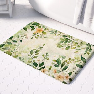 May include: A beige bath mat with a floral pattern featuring green leaves and orange and white flowers. The mat has a textured surface.