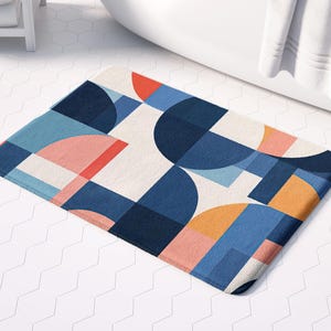 May include: A rectangular bath mat with a geometric design. The pattern features blue, orange, and pink shapes on a white background. The mat is placed on a white tiled floor near a white bathtub.