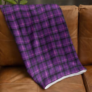 May include: A purple and black plaid blanket with a white underside is draped over a brown leather couch. The blanket features a classic tartan pattern in shades of purple and black, creating a cozy and inviting aesthetic.