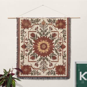 Puede incluir: Un tapiz de pared tejido con un dise&ntilde;o floral geom&eacute;trico. El patr&oacute;n central de estrella est&aacute; en tonos marrones y amarillos, rodeado de hojas verdes y flores rojas sobre un fondo crema. Los bordes tienen un borde con flecos.