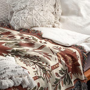 Puede incluir: Una manta crema con un estampado floral en tonos marrones y verdes, bordes con flecos y cojines decorativos. La manta est&aacute; colocada sobre una cama, creando un ambiente acogedor y atractivo.
