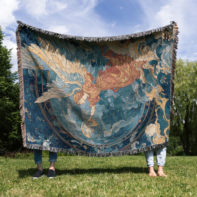 May include: A woven tapestry blanket featuring a celestial design with an angel figure, wings, and a globe. The color palette includes shades of blue, gold, and peach. The blanket has a fringed edge and is displayed outdoors on a sunny day.