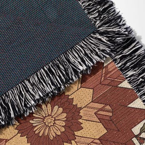 Puede incluir: Primer plano de una manta tejida con un patr&oacute;n floral y geom&eacute;trico. La manta presenta un tejido texturizado en color verde azulado y burdeos, con un borde con flecos en blanco y negro. La parte inferior muestra un dise&ntilde;o floral marr&oacute;n y beige.