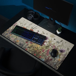 May include: A large, rectangular desk mat with a floral design featuring a variety of flowers and plants in shades of pink, purple, green, and yellow. A black keyboard with rainbow lighting and a black mouse are on the mat.