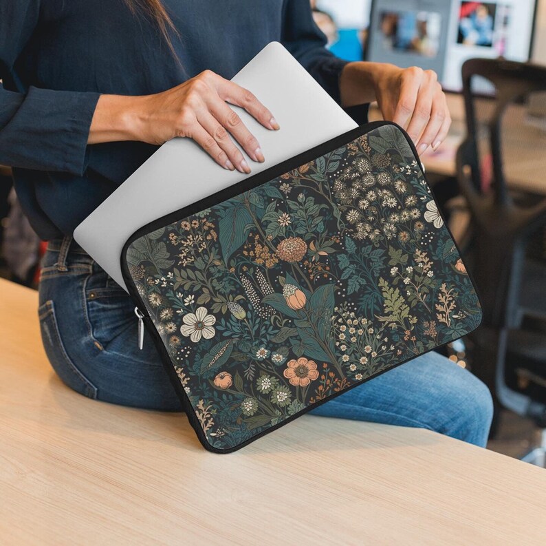 Pode incluir: Uma capa para laptop com um padr&atilde;o floral. A capa &eacute; preta com um design floral verde escuro, branco e laranja. A capa est&aacute; sendo segurada por uma pessoa, mas as m&atilde;os da pessoa n&atilde;o s&atilde;o vis&iacute;veis.