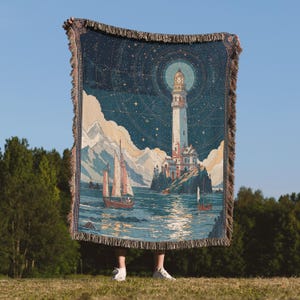May include: A woven throw blanket depicting a lighthouse scene with sailboats, mountains, and a starry night sky. The blanket has a fringed edge and is held up against a blue sky.