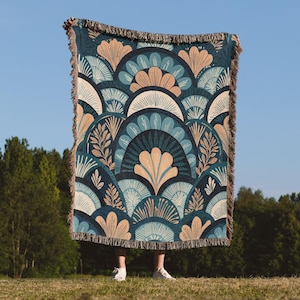 Peut inclure: Une couverture décorative avec un motif répétitif de ventilateurs et de feuilles dans des tons de bleu sarcelle, beige et bleu marine. La couverture a une bordure à franges et est présentée sur fond de ciel bleu.