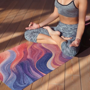 May include: Yoga mat featuring a vibrant, flowing design in red, orange, purple, and blue hues. The mat is rectangular and placed on a wooden floor. A person is seated on the mat in a meditative posture.