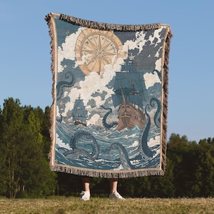May include: A woven blanket featuring a nautical scene with ships, a compass rose, and a giant squid in shades of blue, tan, and cream. The blanket has a fringed edge and is held up against a blue sky.