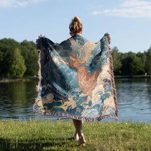 May include: A teal and beige throw blanket with a celestial angel design. The blanket features a detailed illustration of an angel with outstretched wings, clouds, and stars. The blanket has a fringed edge and is held up in front of a person.