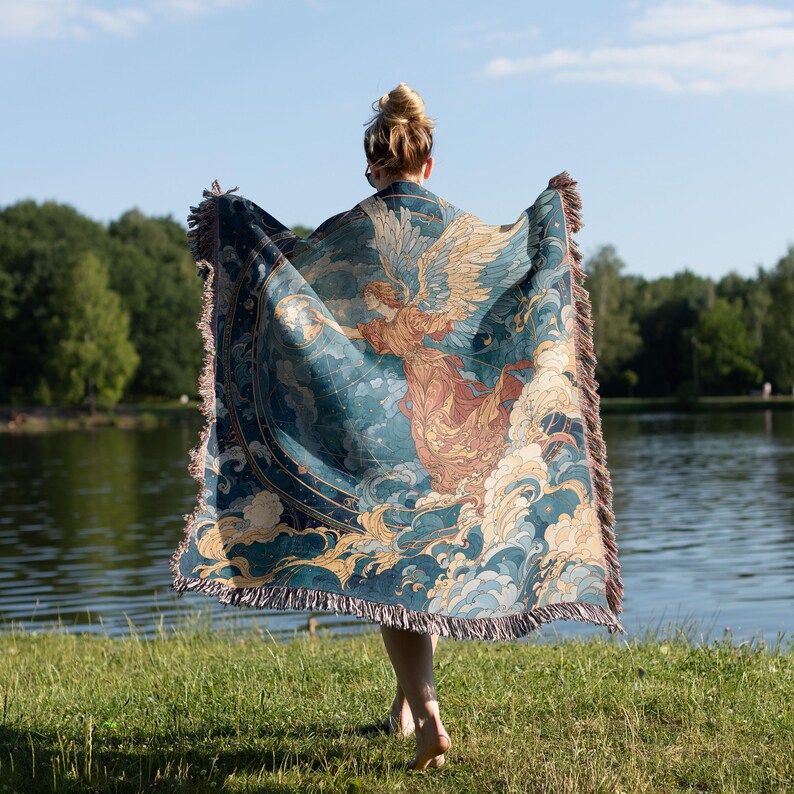 May include: A teal and beige throw blanket with a celestial angel design. The blanket features a detailed illustration of an angel with outstretched wings, clouds, and stars. The blanket has a fringed edge and is held up in front of a person.
