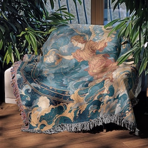 May include: A throw blanket draped over a chair, featuring a celestial design. The blanket showcases an angel figure in shades of peach and gold, reaching towards a globe, set against a backdrop of teal and gold clouds. The blanket has a fringed edge.
