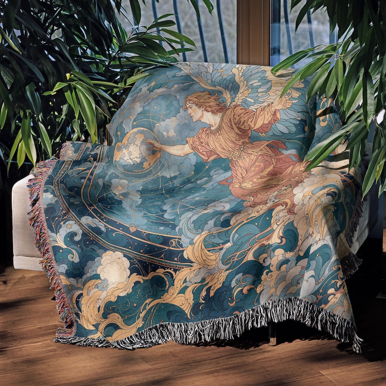 May include: A throw blanket draped over a chair, featuring a celestial design. The blanket showcases an angel figure in shades of peach and gold, reaching towards a globe, set against a backdrop of teal and gold clouds. The blanket has a fringed edge.