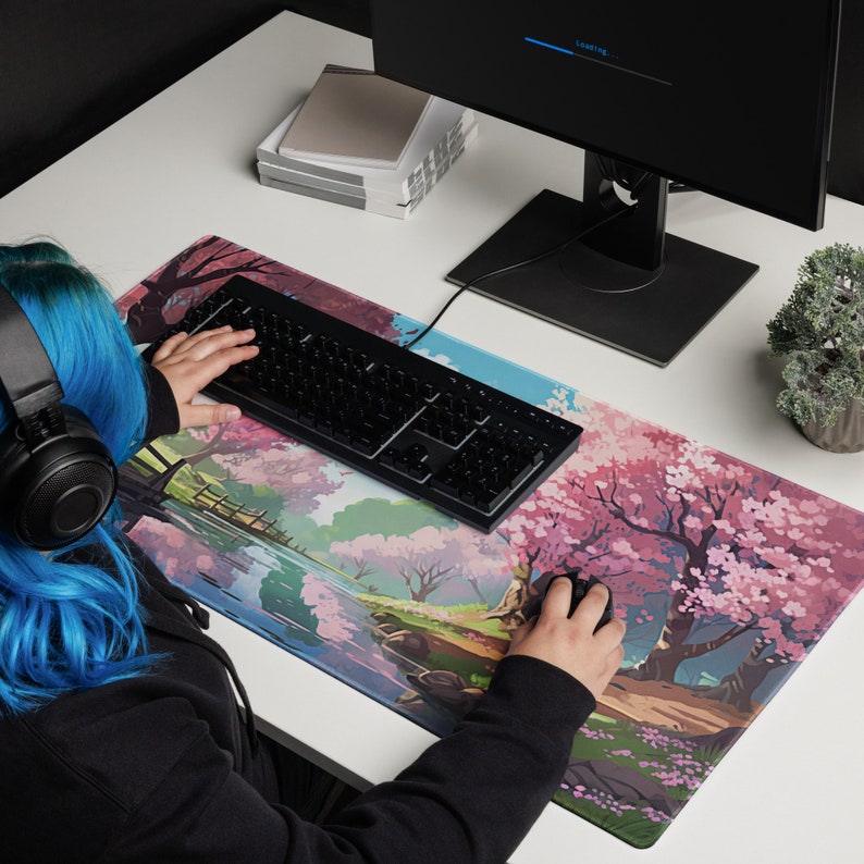 May include: A black keyboard with a white background and a pink and green mousepad with a cherry blossom design. The mousepad has a river and trees in the background.