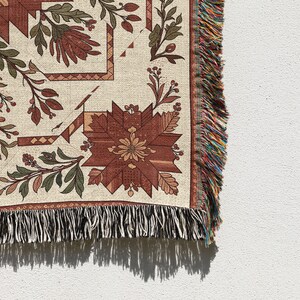 Puede incluir: Una manta tejida con un estampado floral en tonos rojos, verdes y beige. El dise&ntilde;o presenta flores y hojas estilizadas, con un borde con flecos multicolores. La manta se muestra sobre un fondo blanco.