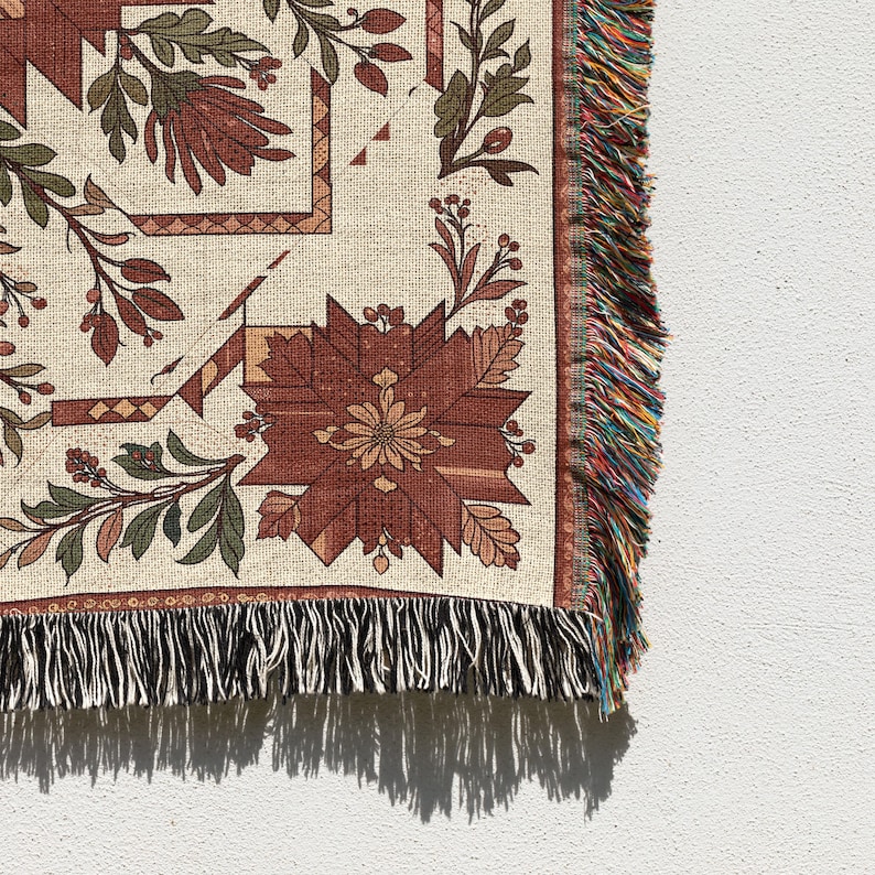 Puede incluir: Una manta tejida con un estampado floral en tonos rojos, verdes y beige. El dise&ntilde;o presenta flores y hojas estilizadas, con un borde con flecos multicolores. La manta se muestra sobre un fondo blanco.